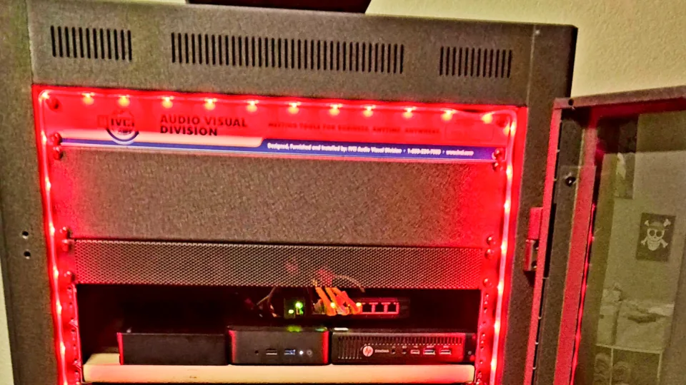 An open server rack lit by red LED strips, holding two compact mini-PC nodes and a red 1U appliance with patch cables, above empty shelves.